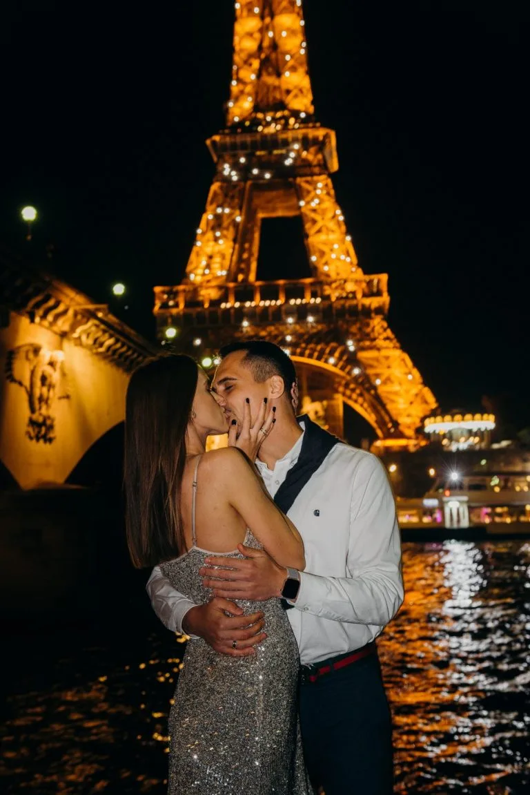 A romantic couple shares a passionate kiss in front of the illuminated Eiffel Tower at night, with the Seine River reflecting the golden lights. The woman wears a sparkling dress, and the man embraces her warmly, capturing the magic of a Paris night photoshoot.